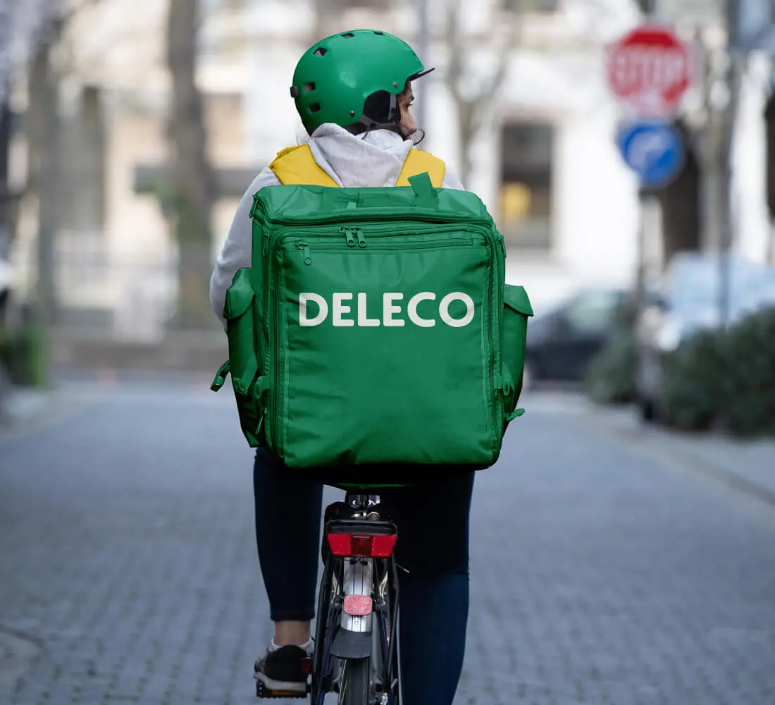 Doručovateľka na bicykli so zelenou taškou, na ktorej je výrazne zobrazené logo Deleco, čím sa zdôrazňuje ekologická doručovacia služba.